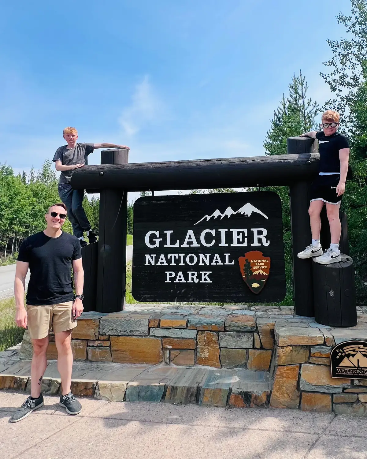 Tim and his two sons at the Glacier National Park entrance sign: one son stands on the stone base at the right, the other leans on the sign's top rail, and Tim stands at the left in a black tee.